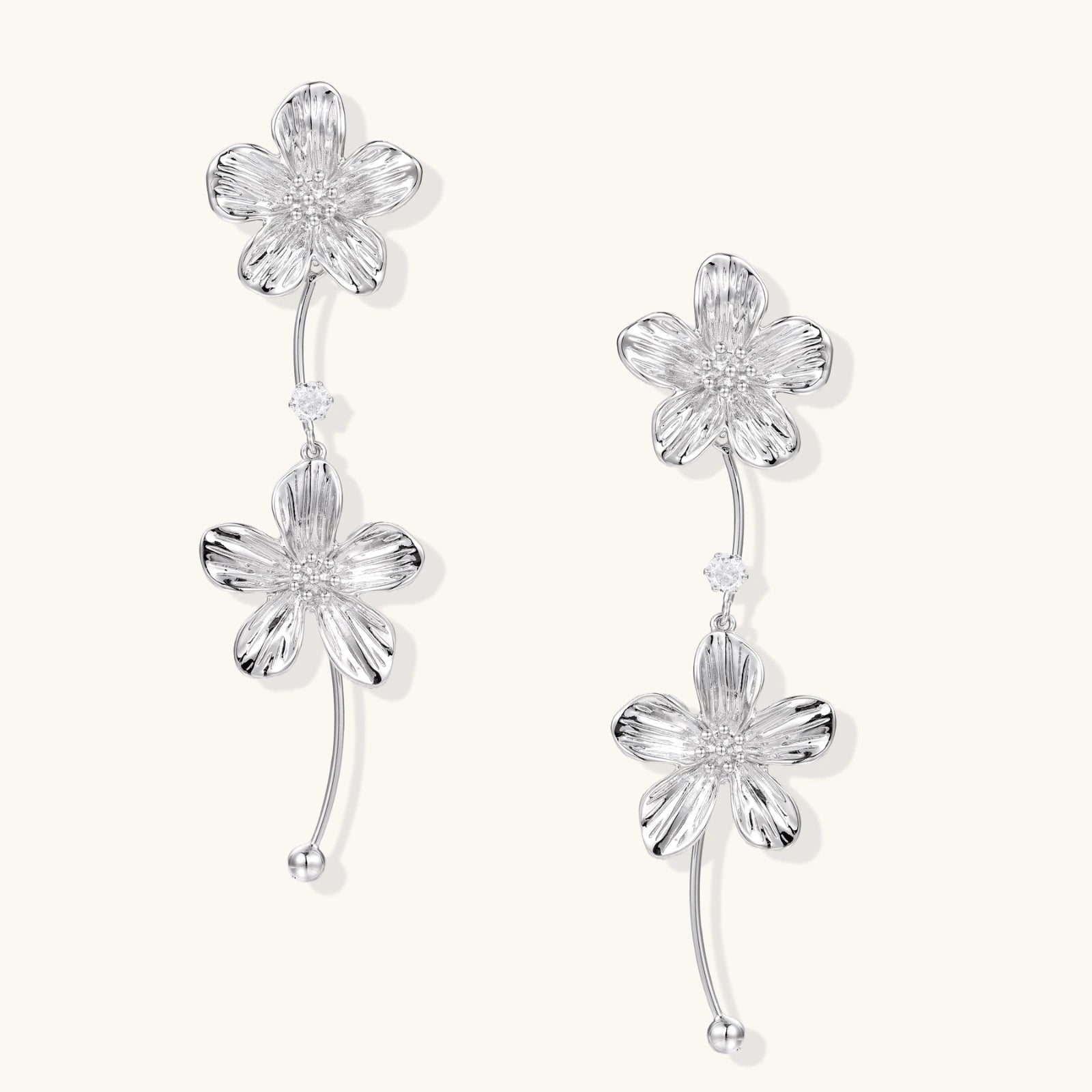 Silver-white asymmetric earrings with textured flower petals and crystal centers on cream background